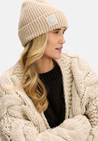 Beżowa prążkowana dzianinowa czapka z małą metką marki. Ciepła, miękka w dotyku. Noszona z kremowym grubym swetrem w warkocze i ciemnym swetrem pod spodem.