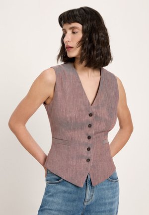 Frau mit schulterlangem dunklem Haar trägt ärmellose, mit Knöpfen versehene mauvefarbene Weste und blaue Jeans, Hände hinter dem Rücken, schaut zur Seite.