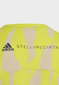 Gul sporttop med beige abstrakta mönster, svart Adidas-logga och "STELLAMCCARTNEY" tryckt på baksidan. Mjuk tygtextur.