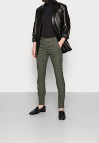 Blazer de cuero negro, cuello alto negro ajustado y pantalones a cuadros verdes. Mocasines negros con ligero brillo, textura suave y corte entallado.