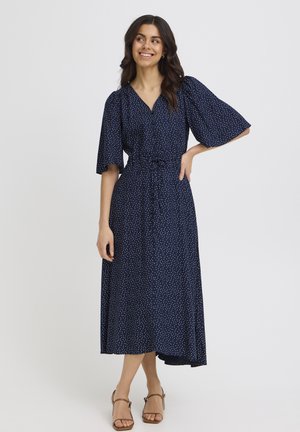 Kobieta uśmiechająca się, ubrana w granatową sukienkę midi w białe grochy, z krótkimi szerokimi rękawami, dekoltem w kształcie litery V i beżowymi sandałami na obcasie.