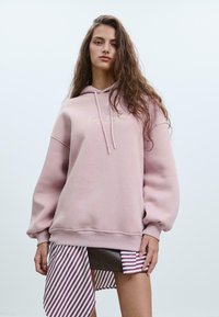 Svetlo ružový oversized mikina s kapucňou a šnúrkou na stiahnutie s rebrovaným lemom. Obsahuje vyšívaný text na hrudi. Pod ňou visí pruhovaná látka.