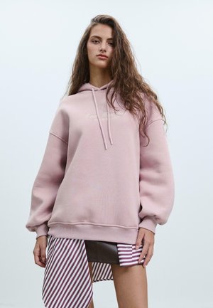 Felpa oversize rosa chiaro con cappuccio regolabile e orlo a coste. Presenta testo ricamato sul petto. Tessuto a righe pende sotto.
