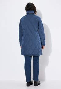 Blå vadderad lång kappa med hög krage, lätt material och diamantmönster. Kombinerad med mörkblå jeans och svarta stövlar.