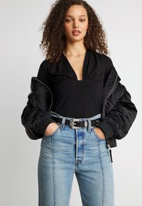 Γυναίκα με σγουρά μαλλιά φορώντας μαύρο τοπ με βαθύ V, μαύρο μπουφάν bomber με τους ώμους ακάλυπτους, ξεβαμμένο μπλε τζιν και μαύρη ζώνη με ασημί αγκράφα.