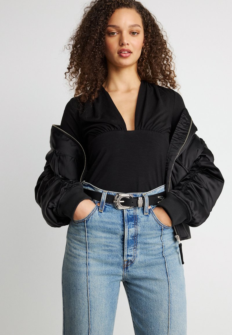 Γυναίκα με σγουρά μαλλιά φορώντας μαύρο τοπ με βαθύ V, μαύρο μπουφάν bomber με τους ώμους ακάλυπτους, ξεβαμμένο μπλε τζιν και μαύρη ζώνη με ασημί αγκράφα.