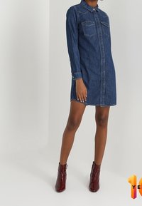 Denim-mekko, jossa on kauluspääntie, pitkät hihat ja kaksi rintataskua. Mekko on syvän sininen ja siinä on suora siluetti.