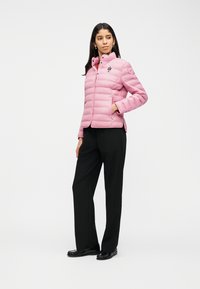 Γυναίκα που φοράει ανοιχτό ροζ καπιτονέ μπουφάν με ψηλό γιακά, μαύρο παντελόνι και μαύρα μοκασίνια, όρθια με το ένα χέρι στην τσέπη.