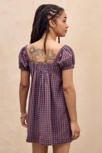 Crvena i plava karirana haljina s napuhanim rukavima, nabrane leđa i otvorenim leđima, koja prikazuje vidljivu tetovažu na lijevom ramenu.