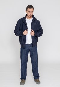 Marineblaue Bomberjacke mit hohem Kragen, Reißverschluss und elastischen Bündchen. Getragen über einem weißen T-Shirt, kombiniert mit blauen Jeans und grauen Sneakers.