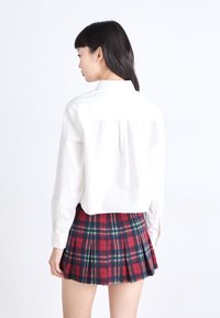 Camisa blanca de cuello con mangas largas, combinada con una falda plisada a cuadros en rojo y azul marino. El conjunto presenta un diseño limpio y patrones distintivos.