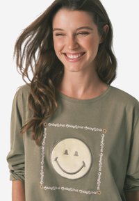 Oliwkowa koszulka z długim rękawem z dużą grafiką uśmiechniętej twarzy, otoczoną napisem "SmileyWorld." Wykonana z miękkiej bawełny.