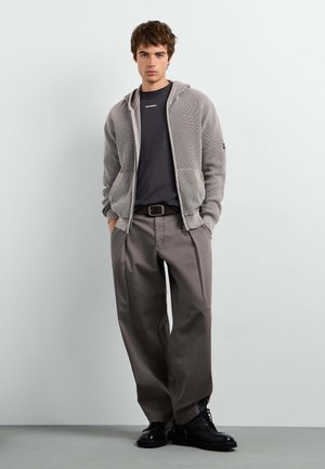 Giovane uomo in piedi contro un muro semplice che indossa una felpa grigia con cappuccio e zip, una t-shirt nera, pantaloni grigi larghi, cintura nera e stivali neri.