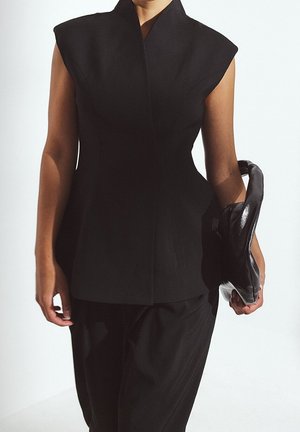 Mujer con un top negro sin mangas estructurado y pantalones negros, sosteniendo un gran bolso de mano de cuero negro a su lado.