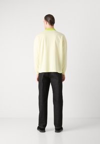 Camisa amarilla claro de manga larga con un cuello verde, combinada con pantalones de mezclilla oscuros y zapatos negros, vista desde la parte de atrás.