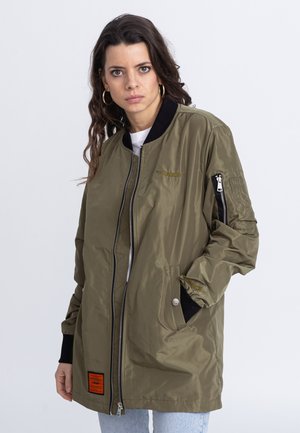 Veste bomber vert olive, tissu brillant, col noir côtelé, poche latérale zippée, étiquette orange en bas, coupe décontractée, détails de couture visibles.