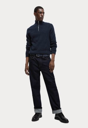 Námořnický svetr s polovičním zipem, žebrovaná textura, spárovaný s tmavými džíny s ohrnutými konci, doplněný černými šněrovacími botami a modrým opaskem.