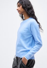 Ανοιχτό μπλε πουλόβερ με απαλό ύφασμα, στρογγυλή λαιμόκοψη και ριμπ μανσέτες. Φορεμένο με μαύρο, ψηλόμεσο παντελόνι που δείχνει άνετη εφαρμογή.