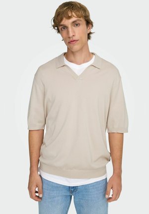 Młody mężczyzna w beżowej, krótkim rękawie, dzianinowej koszulce polo na białym t-shircie i jasnoniebieskich dżinsach na tle jednolitej, białej ściany.