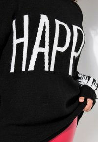 Schwarzer Pullover mit fettem, weißen „HAPPY“-Text. Besitzt eine weiche Textur und gemusterte Schwarz-Weiß-Bündchen. Insgesamt lockere Passform.