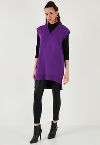 Lila kötött ujjatlan felső V-nyakkal, bordás alsó résszel, fekete leggings-szel és bokacsizmával párosítva. Az anyag sima tapintású.