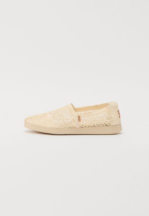 Beige slip-on-sko med detaljeret blomsterblonde-design og flad sål, sidevisning på hvid baggrund.