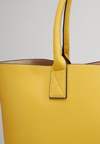 Gros plan d'un sac fourre-tout en cuir jaune montrant la poignée et le bord supérieur avec des coutures détaillées.