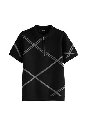 Junge Person mit geflochtenem Haar trägt ein schwarzes Polo-Shirt mit kurzen Ärmeln, grauen diagonalen Streifen und einem Frontreißverschluss, Hände in den Taschen.