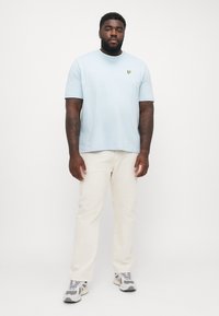 Lyseblå bomulds T-shirt med et lille gult logo, parret med cremefarvede bukser og grå sneakers med sorte detaljer.