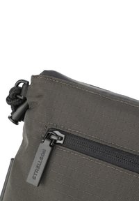 Borsa di tessuto grigia con una texture liscia, dotata di una tasca frontale con zip e una chiusura a clip, decorata con un'etichetta logo nera.