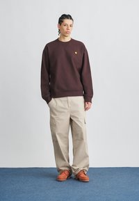 Felpa color marrone con polsini a coste, pantaloni cargo color crema con tasche laterali e scarpe arancioni con suola scura, su sfondo grigio.