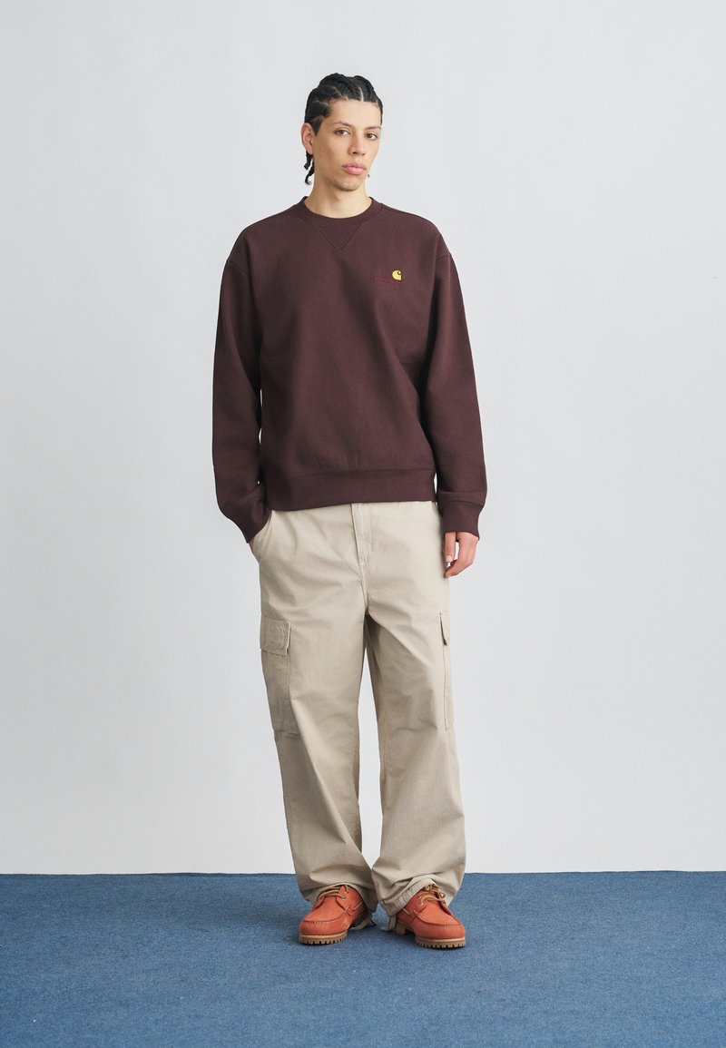 Bordeauxrotes Sweatshirt mit gerippten Bündchen, cremefarbene Cargohose mit Seitentaschen und orangefarbene Schuhe mit dunklen Sohlen vor grauem Hintergrund.