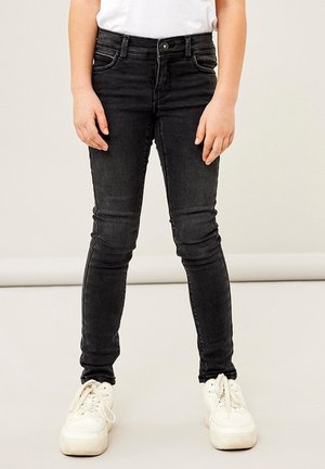 Vaqueros skinny negros de textura suave, con cinco bolsillos y cierre de botón, combinados con zapatillas blancas.