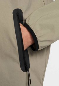 Beige jacka med svarta detaljer, med en dragkedjeficka på ärmen och en elastisk manschett. Mjuk, slät tyg med matt finish.