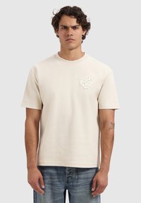 Lichtbeige katoenen T-shirt met korte mouwen, met een textuurlogo op de borst. Zachte textuur en een relaxte pasvorm. In combinatie met blauwe spijkerbroek.