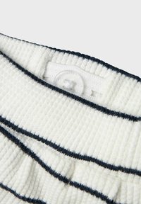 Tessuto bianco testurizzato con strisce orizzontali blu navy. Presenta un bottone trasparente tondo e un colletto a coste. Visto ravvicinato dei dettagli dell'indumento.
