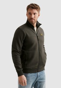 Hombre con una chaqueta acolchada color verde oscuro con cremallera y un parche en el pecho, y vaqueros azul claro, mirando hacia un lado.