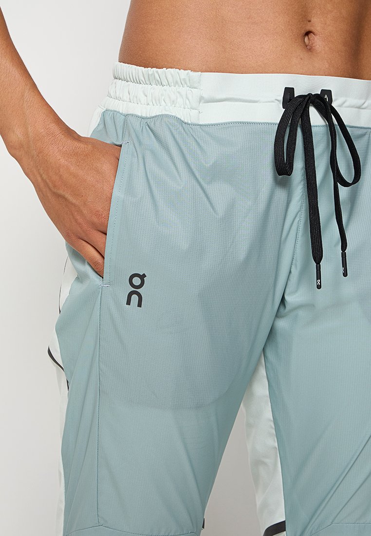 Männlicher Oberkörper, der hellblaue Sportshorts mit schwarzem Kordelzug trägt, die Hand in der Tasche, und das sichtbare "On"-Markenlogo am linken Oberschenkel.