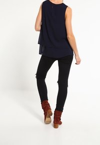 Blouse sans manches bleu marine avec un design en couches, associée à un jean skinny noir avec des détails déchirés, et des bottines marron à talons.