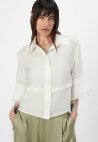 Blouse blanche légère avec des détails à franges aux épaules et à l'ourlet. Dotée d'un col et de manches mi-longues. Tissu texturé.