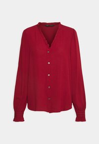 Blouse rouge à manches longues avec col en V volanté, boutonnage sur le devant et poignets froncés sur un fond clair uni.