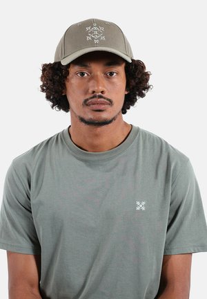 Homme aux cheveux bouclés portant une casquette gris-vert et un t-shirt assorti avec un petit logo blanc brodé sur le côté gauche de la poitrine, sur un fond uni.