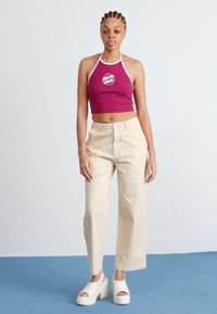 Burgundská crop top s bielym lemovaním a logom, spárovaná so smotanovými širokými nohavicami a bielymi platformovými drevákmi na modrej podlahe.