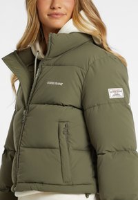 Alyvuogių žalia pūkinė striukė su užtrauktuku priekyje, aukštu apykakle ir logotipu. Pasitikti su pagalvėles tekstūra, šoninėmis kišenėmis ir reguliuojama kapuce.