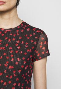 Gros plan d'une personne portant un haut noir transparent à manches courtes avec un motif floral rouge sur un fond clair uni.