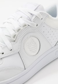 Vit sneaker i slät läder med perforerade detaljer, platta vita snörningar och en rund präglad logotyp på sidan. Texturerad sula.