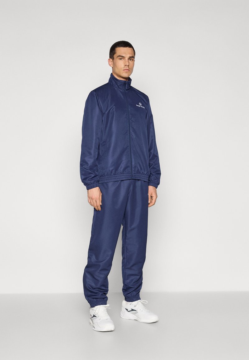 Marinefarbener Trainingsanzug aus glattem Stoff, bestehend aus einer Reißverschlussjacke mit hohem Kragen und elastischen Bündchen, kombiniert mit passenden Hosen, getragen mit weißen Sneakers.