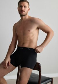 Schwarze Boxer-Shorts aus glattem Material mit einem markierten Bund, die ein enganliegendes Design aufweisen. Das Modell sitzt auf einem Holzstuhl.