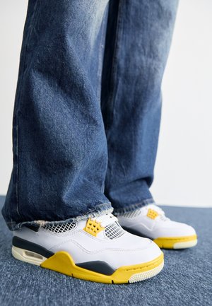 Sneakers bianchi e gialli con una finitura testurizzata, dettagli in rete e un colletto imbottito, abbinati a jeans di denim blu strappati.