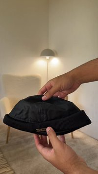 Chapéu tipo balde preto com aba em malha canelada. A superfície é lisa e brilhante, com uma pequena etiqueta dourada com o logótipo na lateral.